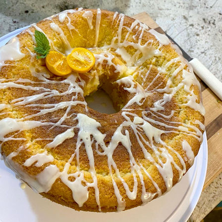 Glazed lemon bundt cake with white icing drizzle, kumquat halves and mint garnish, one slice removed, on a white plate atop a wooden cutting board