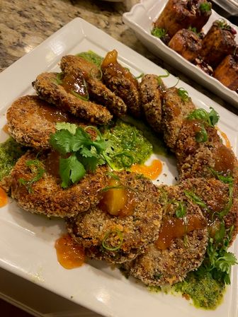 Crispy panko-crusted round fritters arranged on a white square plate over green herb sauce, drizzled with orange chutney and garnished with cilantro and sliced green onions.