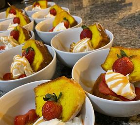 Rows of individual dessert bowls with golden pound cake slices, fresh strawberries, whipped cream and caramel drizzle on a granite countertop