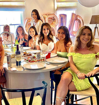 Smiling women gathered around a bright kitchen island enjoying a bridal shower brunch with wine, plated food and rose-gold 'BRIDE' balloons.