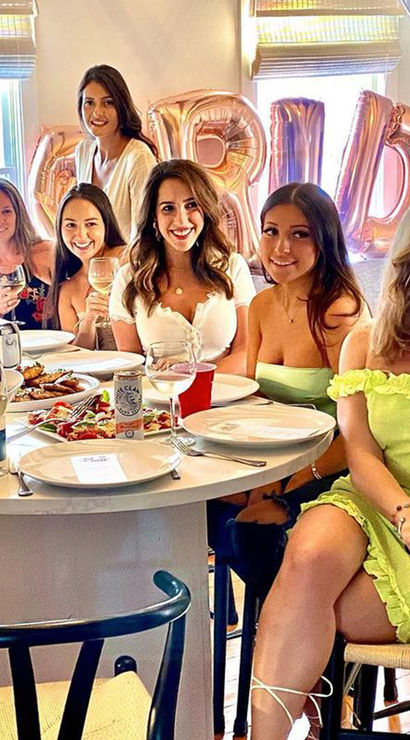 Smiling women gathered around a bright kitchen island enjoying a bridal shower brunch with wine, plated food and rose-gold 'BRIDE' balloons.