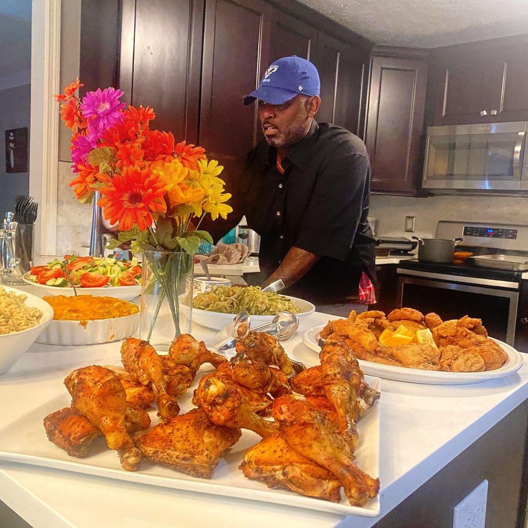 Home kitchen buffet-style dinner spread with a colorful bouquet, a white platter of golden roasted chicken drumsticks and thighs in the foreground, plates of crispy fried chicken, green beans, salad, rice and casserole, and a man in a blue cap preparing food at the island.