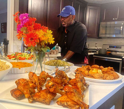 Home kitchen buffet-style dinner spread with a colorful bouquet, a white platter of golden roasted chicken drumsticks and thighs in the foreground, plates of crispy fried chicken, green beans, salad, rice and casserole, and a man in a blue cap preparing food at the island.