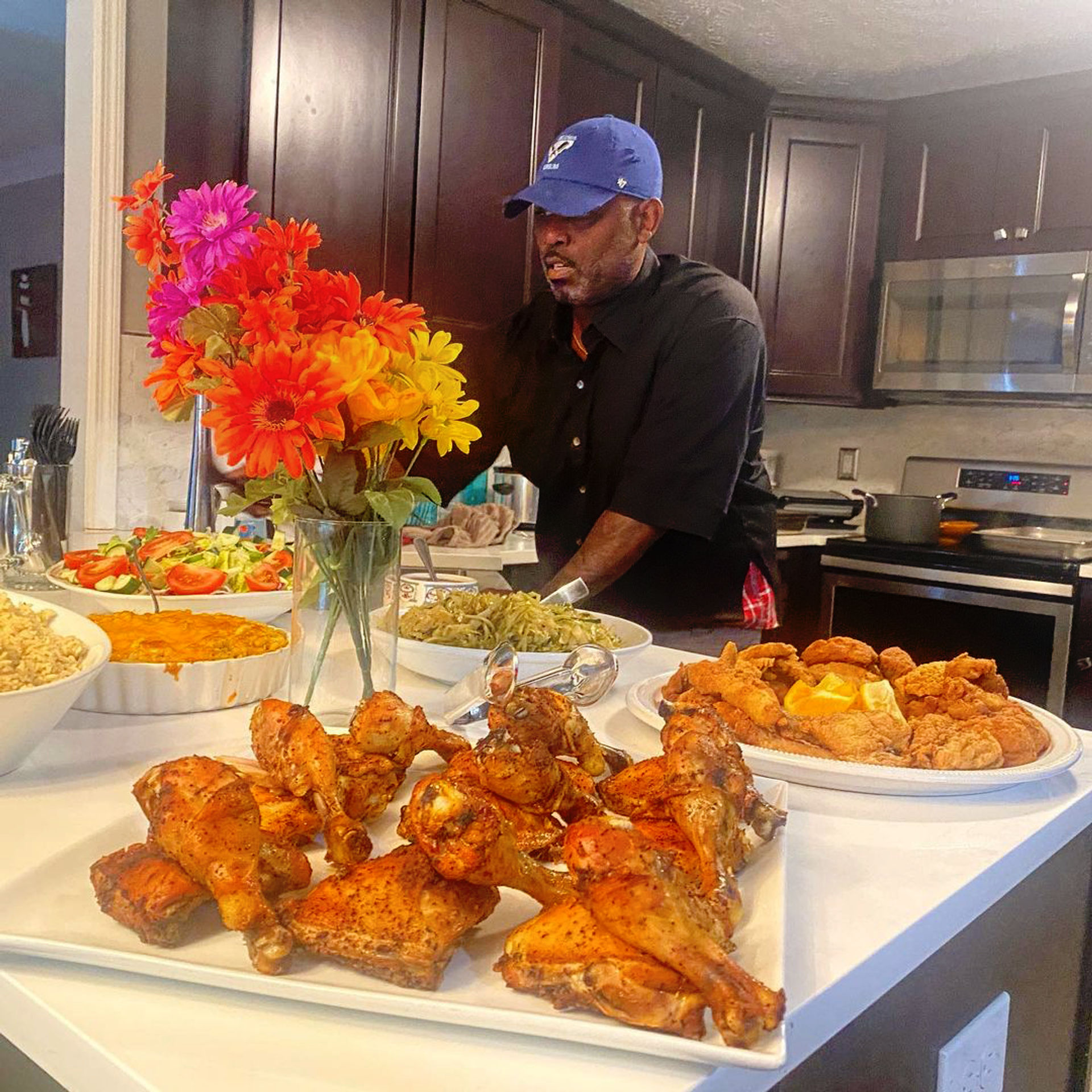 Home kitchen buffet-style dinner spread with a colorful bouquet, a white platter of golden roasted chicken drumsticks and thighs in the foreground, plates of crispy fried chicken, green beans, salad, rice and casserole, and a man in a blue cap preparing food at the island.