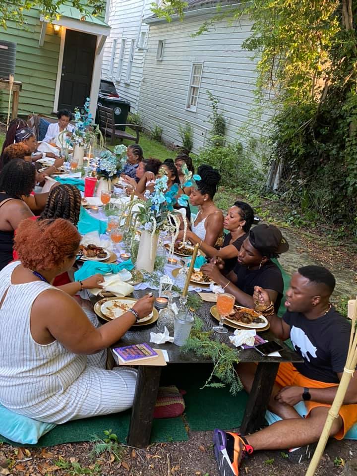 Backyard al fresco dinner party with guests seated on cushions around a low wooden table, turquoise table accents, floral centerpieces, plates of food and drinks amid garden greenery.