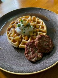 Brunch plate with a golden waffle topped with diced apples, a dollop of whipped cream, chopped herbs and syrup drizzle, served with two browned sausage patties on a dark round plate over a wooden table.
