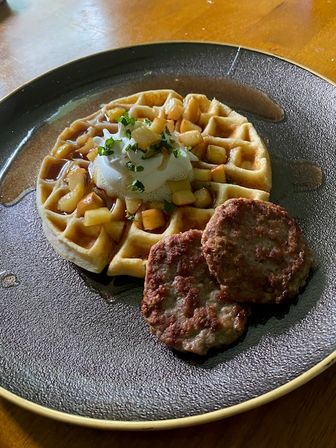 Brunch plate with a golden waffle topped with diced apples, a dollop of whipped cream, chopped herbs and syrup drizzle, served with two browned sausage patties on a dark round plate over a wooden table.