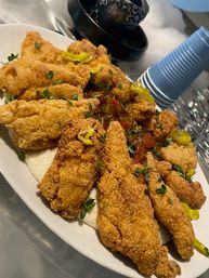Crispy southern-style fried fish fillets on a white plate over creamy grits, topped with pickled peppers, tomato relish and fresh herb garnish, party-style with stacked blue disposable cups in the background