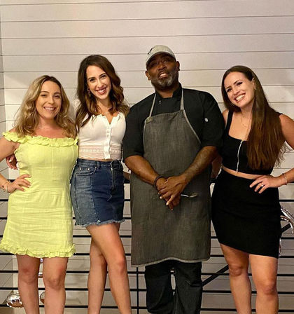Group photo at a casual dining spot: three smiling women in summer outfits posing with a man in a chef’s apron and cap in front of a white shiplap wall.
