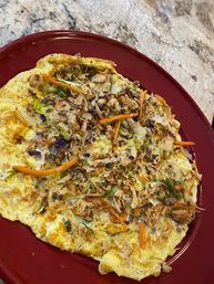 Hearty homemade vegetable omelette with shredded carrots, cabbage and browned bits on a red plate atop a granite kitchen countertop