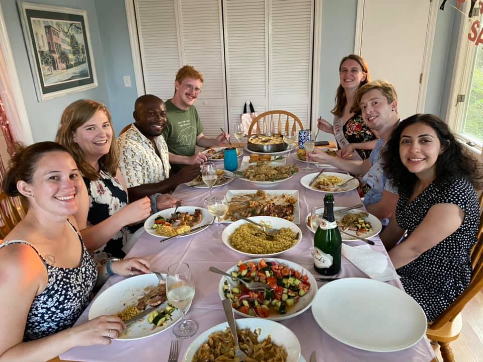 Eight smiling people gathered around a home dining table for a casual dinner party with bowls of rice, roasted vegetables, fresh salad, wine glasses and a champagne bottle in a bright dining room
