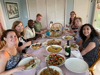 Eight smiling people gathered around a home dining table for a casual dinner party with bowls of rice, roasted vegetables, fresh salad, wine glasses and a champagne bottle in a bright dining room