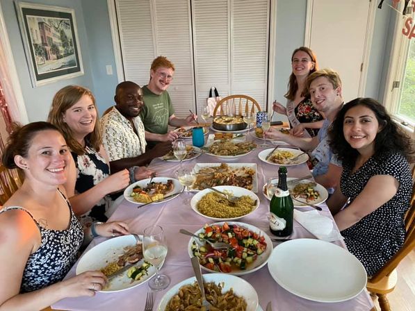 Eight smiling people gathered around a home dining table for a casual dinner party with bowls of rice, roasted vegetables, fresh salad, wine glasses and a champagne bottle in a bright dining room