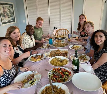 Eight smiling people gathered around a home dining table for a casual dinner party with bowls of rice, roasted vegetables, fresh salad, wine glasses and a champagne bottle in a bright dining room