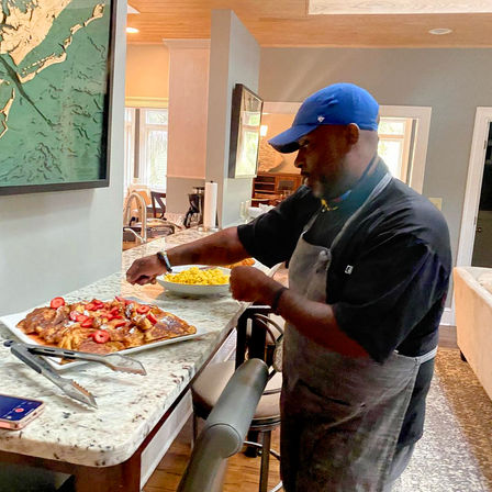 Home cook in a blue cap and apron plating strawberry-topped French toast with a bowl of scrambled eggs on a granite kitchen island in a bright modern home kitchen.