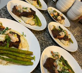 Surf-and-turf plates: seared steak topped with garlic shrimp, whipped mashed potatoes and roasted asparagus on white oval plates lined up on a granite kitchen counter.