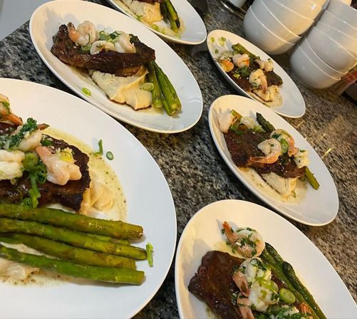 Surf-and-turf plates: seared steak topped with garlic shrimp, whipped mashed potatoes and roasted asparagus on white oval plates lined up on a granite kitchen counter.