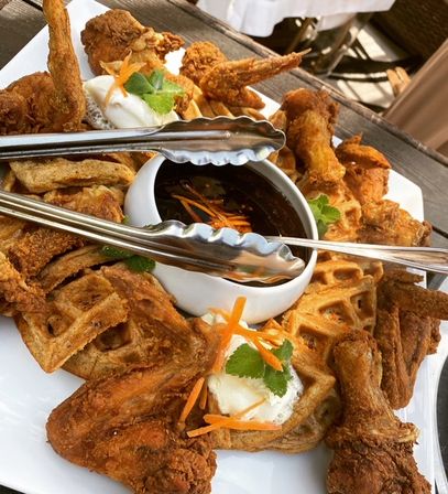 Crispy fried chicken and golden waffles on a white platter with dark dipping sauce, whipped butter, carrot garnish and serving tongs