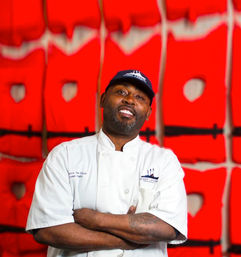 Smiling chef in a white coat and navy cap with arms crossed and a tattooed forearm, posing in front of a bold red abstract wall — upbeat restaurant portrait