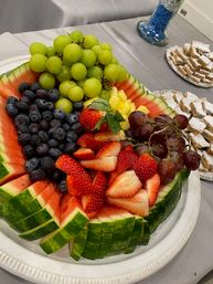 Vibrant fresh fruit platter carved into a watermelon bowl with green and red grapes, blueberries, sliced strawberries and pineapple on a white platter at a catered event table.