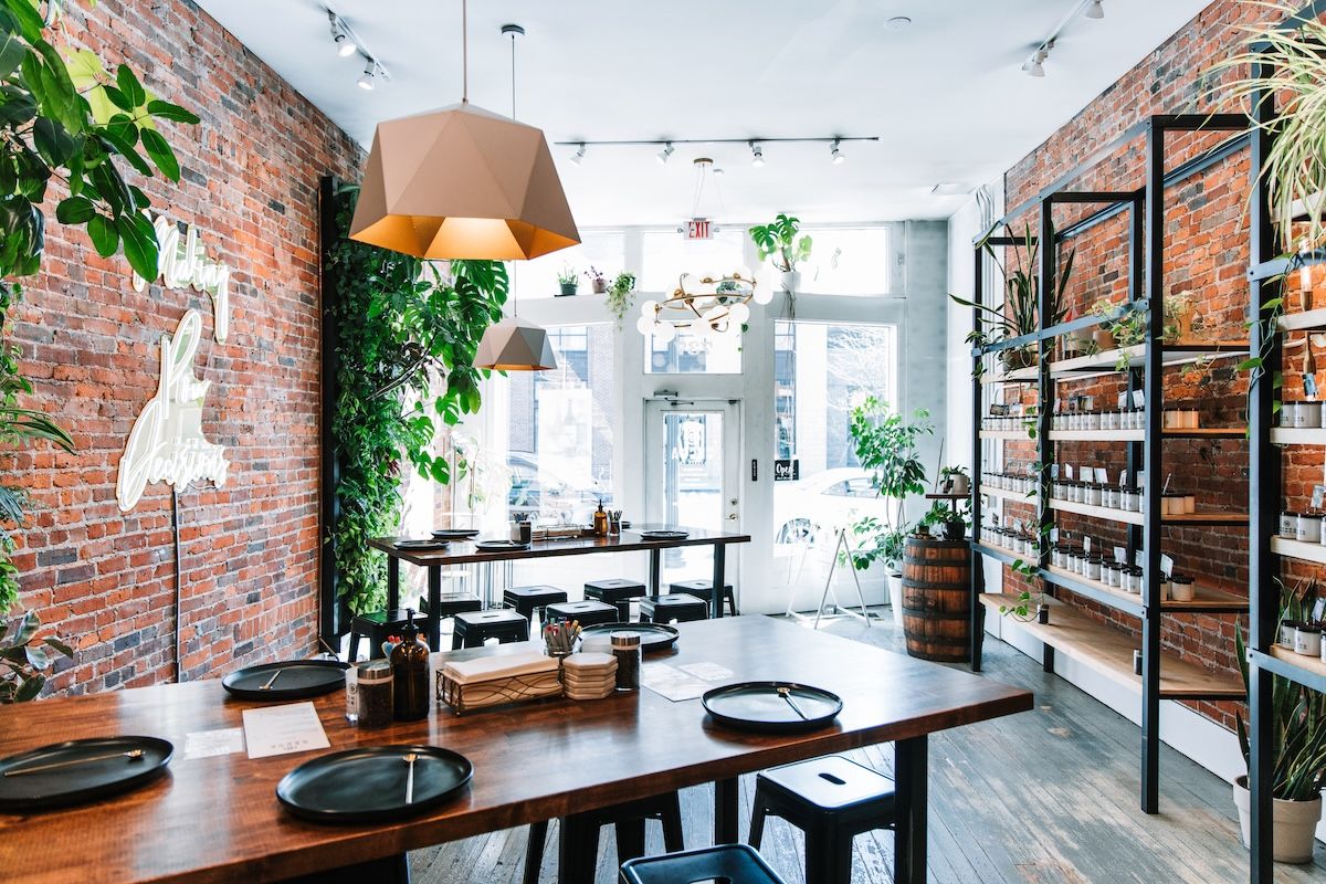 Bright, plant-filled urban cafe interior with exposed brick walls, geometric pendant lamps, communal wooden tables set with black plates, and retail shelving.