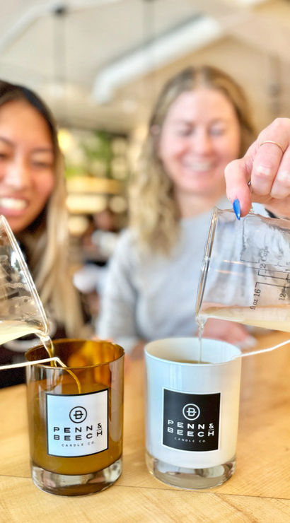 Close-up of two people pouring melted wax from glass measuring beakers into amber and white logo-labeled candle jars on a wooden table during a bright, hands-on indoor candle-making workshop.