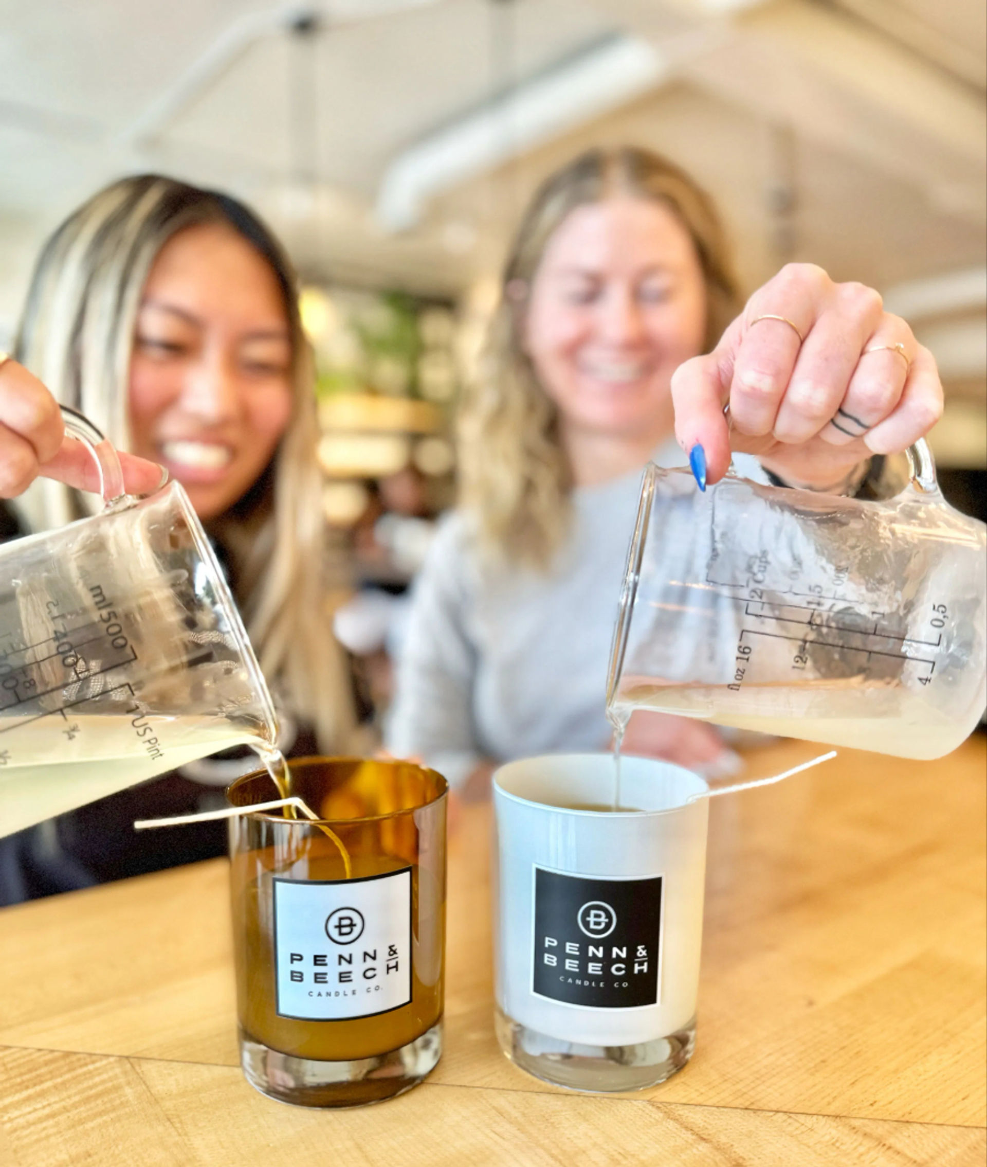 Close-up of two people pouring melted wax from glass measuring beakers into amber and white logo-labeled candle jars on a wooden table during a bright, hands-on indoor candle-making workshop.