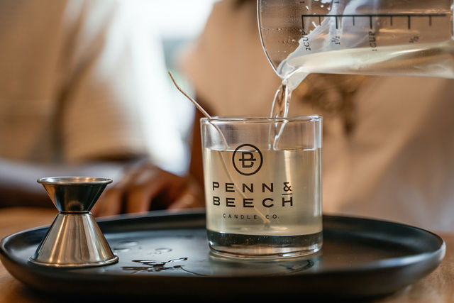 Close-up of pouring warm wax into a clear glass candle jar on a black tray with a metal jigger and stirring spoon — candle-making scene.