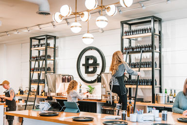 Modern bright coffee shop interior with baristas behind a wooden counter, espresso machines, tall black shelving stocked with bottles reached by a rolling ladder, a large circular wall logo and globe pendant lights.