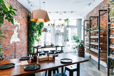 Bright loft‑style urban cafe interior with exposed brick walls, geometric pendant lights, lots of plants, communal wooden tables set with black plates, shelves of jars, and sunlit storefront windows.