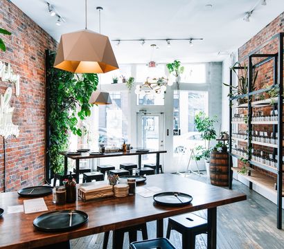 Sunlit urban café interior — industrial-chic exposed brick, geometric pendant lights, long wooden communal tables with black plates and metal stools, lush plants and retail shelves by a street-facing storefront.
