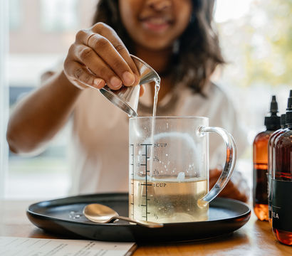 Hand pouring liquid from a metal jigger into a glass measuring cup on a black tray, with amber dropper bottles and a spoon nearby — DIY skincare or herbal remedy mixing in a kitchen.