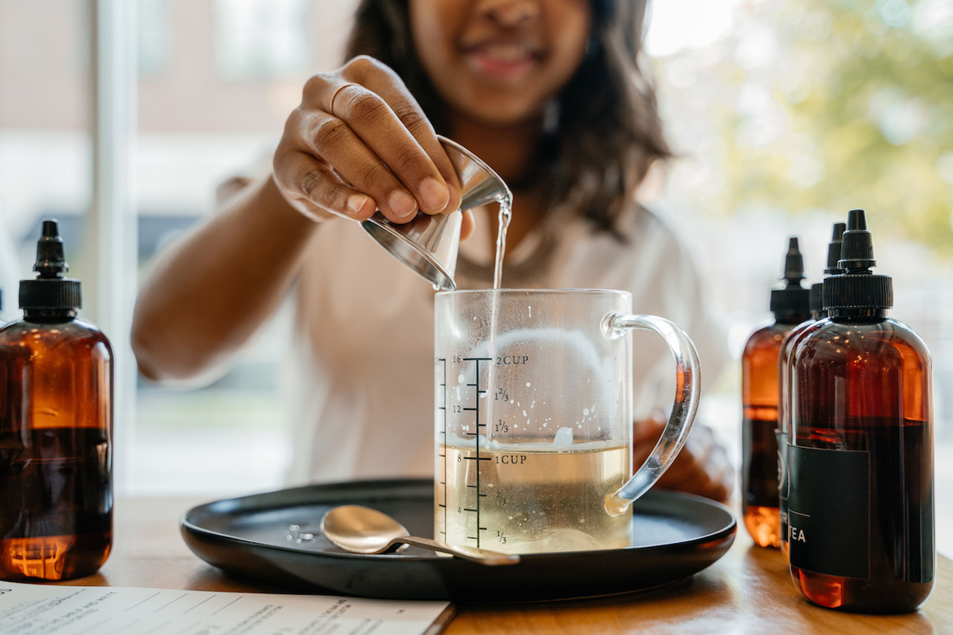 Hand pouring liquid from a metal jigger into a glass measuring cup on a black tray, with amber dropper bottles and a spoon nearby — DIY skincare or herbal remedy mixing in a kitchen.