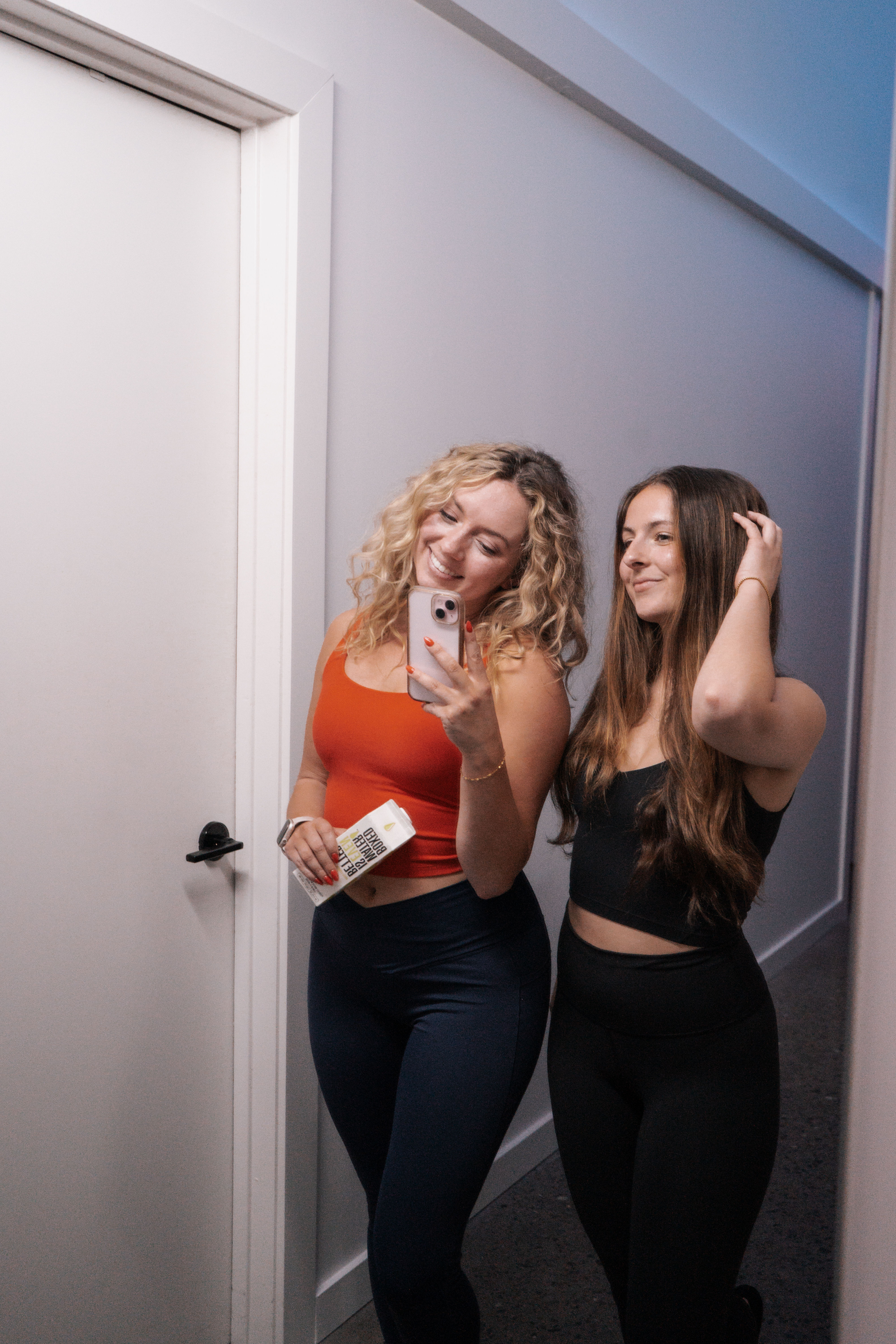 Mirror selfie of two women in a home hallway wearing athleisure—one in an orange crop top holding a phone and a boxed drink, the other in a black crop top and leggings adjusting her hair.