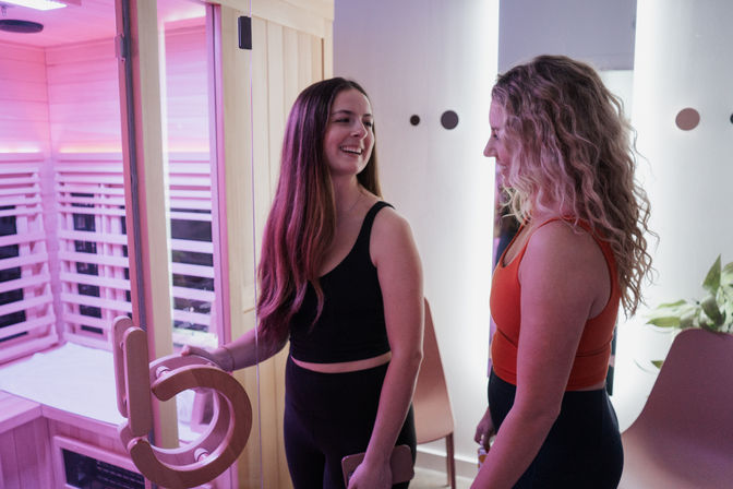 Two women in workout clothes smiling as one opens a pink-lit infrared sauna door in a modern wellness studio