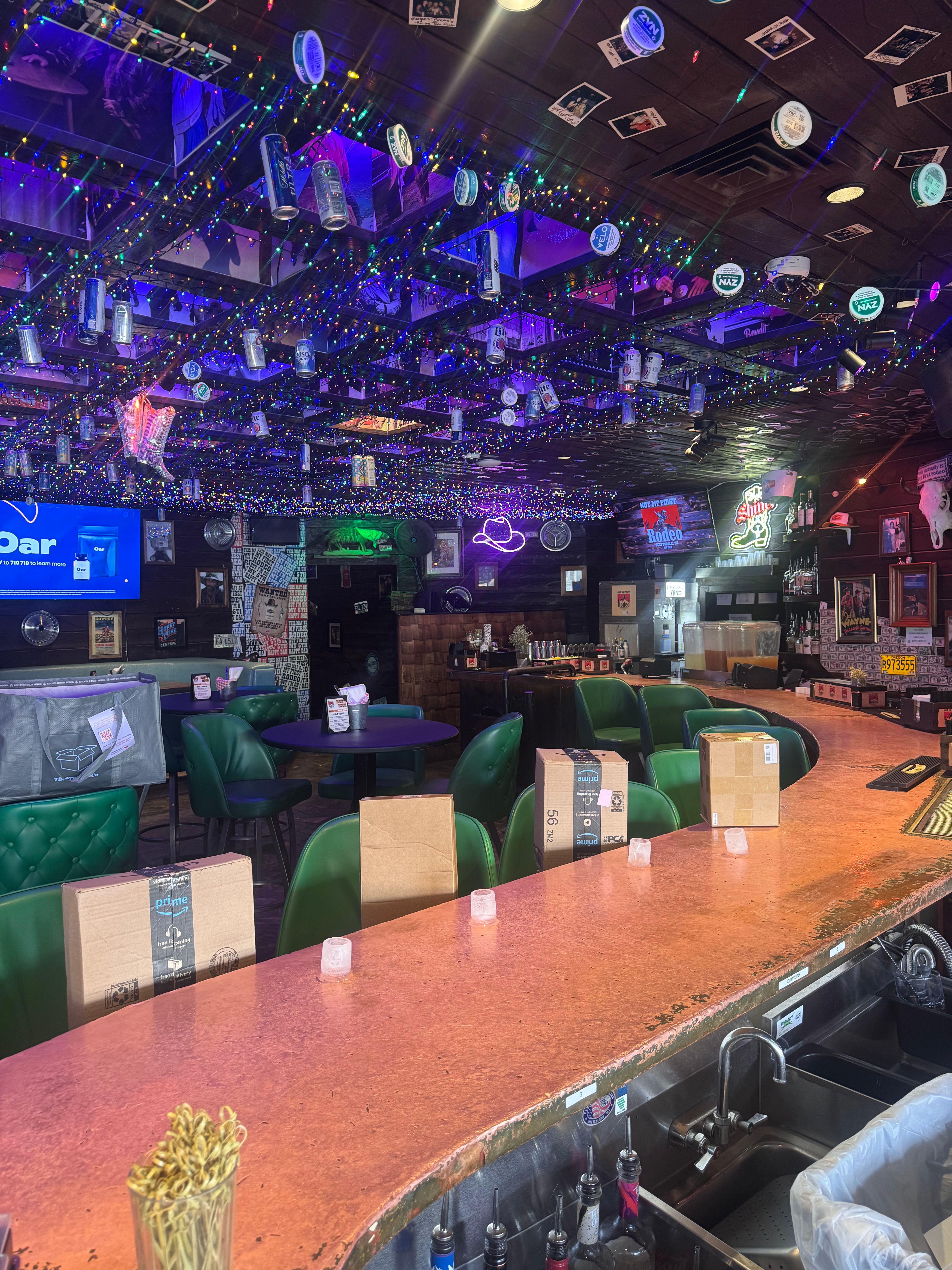 Cozy neighborhood bar interior with a copper-topped counter, green vinyl stools, delivery boxes on seats, neon signs and TVs, and a ceiling draped in multicolored string lights, hanging beer cans and photos.