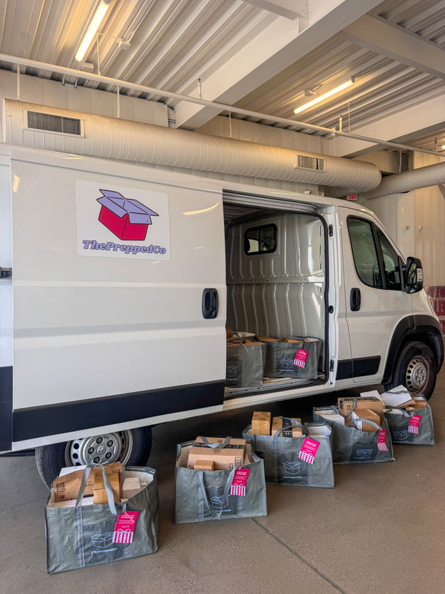 White cargo van in an indoor loading bay with sliding door open and rows of gray reusable delivery bags filled with cardboard packages lined on the concrete, ready for delivery.