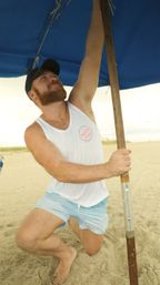 Bearded man in a cap kneeling on a sandy beach, smiling as he secures a blue beach umbrella pole while wearing a white tank top and light-blue swim trunks