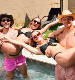 Four friends laughing at a backyard pool party as two men in hats lift two women in black bikinis over the water on a sunny day, sunglasses and fun vibes.