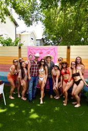 Group of friends in swimsuits and cowboy hats posing on artificial turf in a colorful residential backyard with a pink banner and rainbow wood fence under leafy trees — summer backyard party vibe.