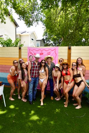 Group of friends in swimsuits and cowboy hats posing on artificial turf in a colorful residential backyard with a pink banner and rainbow wood fence under leafy trees — summer backyard party vibe.