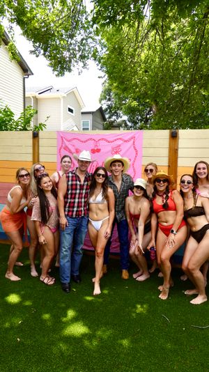 Group of friends in swimsuits and cowboy hats posing on artificial turf in a colorful residential backyard with a pink banner and rainbow wood fence under leafy trees — summer backyard party vibe.