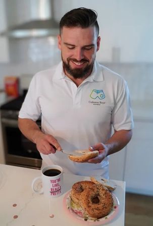Person spreading cream cheese on a toasted everything bagel at a bright modern kitchen counter, with a plate of bagels and a coffee mug reading 'You're like, really pretty' nearby — casual breakfast scene.
