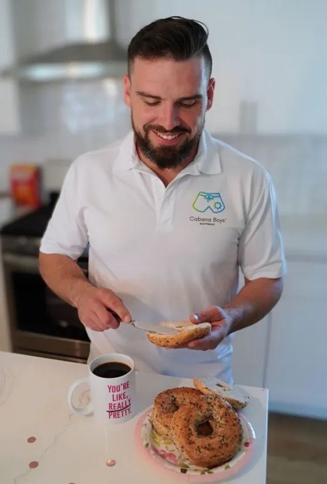 Smiling man in a modern kitchen spreading cream cheese on a sesame bagel from a plate of bagels, with a coffee mug that reads 'YOU'RE LIKE, REALLY PRETTY' — casual breakfast scene.