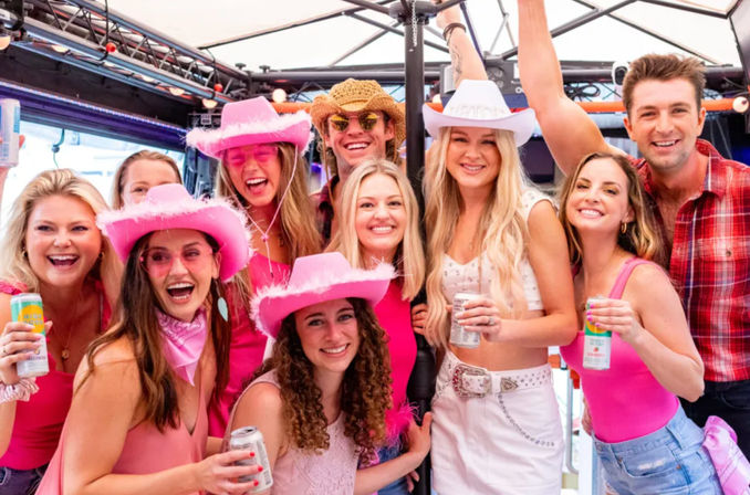 Smiling group of friends in pink cowgirl hats and bright outfits holding canned drinks on a covered party deck, posing for a festive group photo.