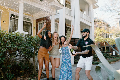 Group of friends celebrating outdoors as a man pops a champagne bottle, spraying foam over three women on the sidewalk in front of a two-story house with white columns in a leafy residential neighborhood.
