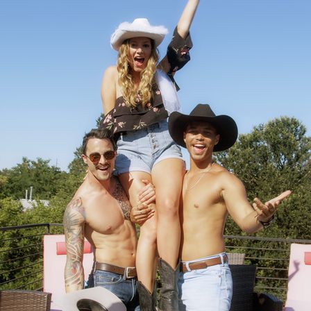 Three friends at a sunny rooftop party: two shirtless men in cowboy hats lift a smiling woman wearing a white cowboy hat, floral top, denim shorts and cowboy boots against a blue sky and treeline.