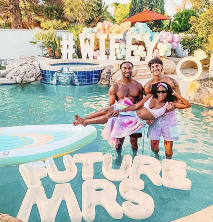 Three people posing in a backyard pool at a pastel balloon bridal party with a large “#WIFEY” sign and floating “FUTURE MRS” letters