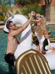 Couple enjoying a backyard pool party on a gold-and-white inflatable unicorn — man feeds a slice while woman in heart-shaped sunglasses holds a champagne bottle.