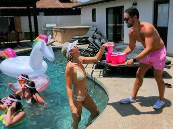 Backyard pool party scene with a shirtless man in pink shorts handing a tray of pink plastic cups to a woman in a white bikini and visor, friends floating on an inflatable unicorn and rings in the sunlit pool.